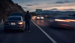 A man stands beside a disabled car with its hood up on a dark highway shoulder near the Camino Del Rio exit, as traffic speeds past with motion-blurred headlights.