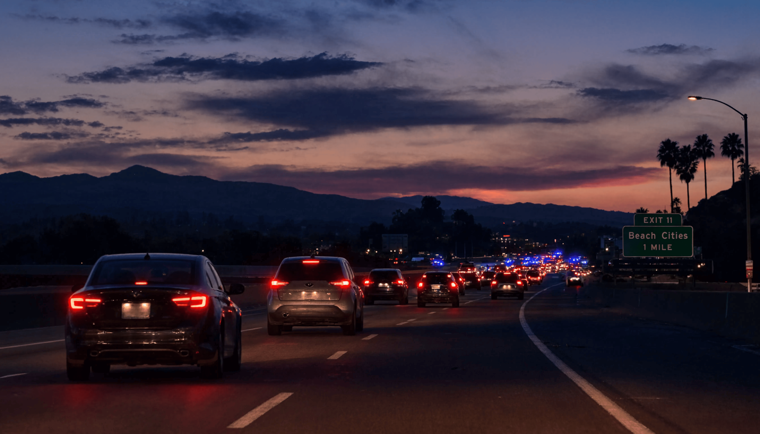 Evening freeway traffic approaching an incident, with police lights flashing ahead, a dramatic sunset sky, and a 'Beach Cities 1 Mile' exit sign on the right.