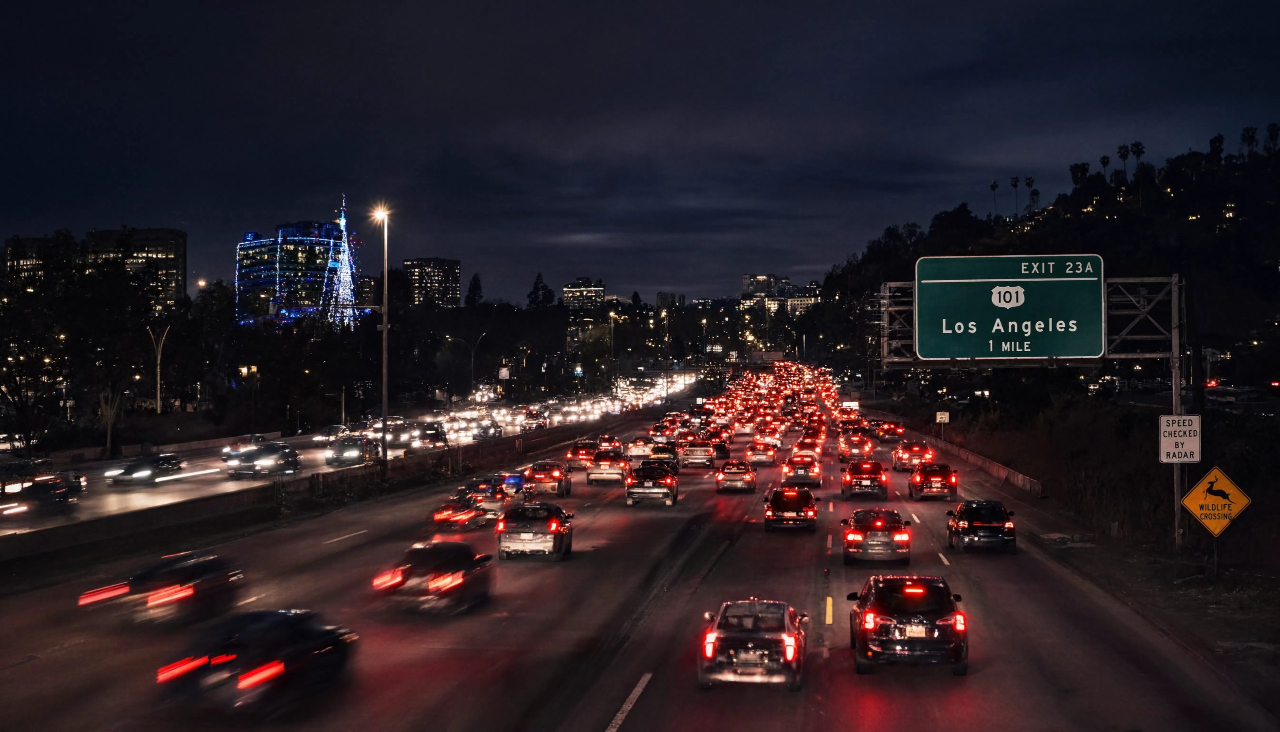 Heavy nighttime traffic on the 101 freeway toward Los Angeles with holiday lights on buildings in the distance